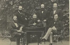 Black and white photo of six bearded men gathered around a table dressed in formal suits from the 1800s, threee seated and three standing